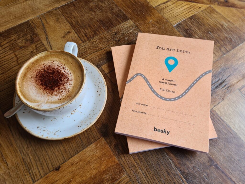 Photo showing a cappuccino on the left with two travel journals 'You are here' on the right hand side. All items are on a wooden table.