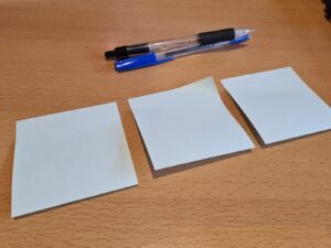 a photo of 3 sticky notes that are pale blue on top of a desk. There are two pens above the sticky notes.