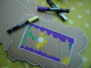 Cardboard photo frame on a table with a yellow spotty tablecloth. The cardboard craft frame is decorated with paint pens and has a flower and purple scallops along the sides.