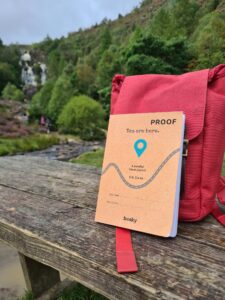 A copy of You are here. travel journal leaning against a red backpack. Both items are on a wooden bench. In the background is a hill covered with forest and a waterfall.